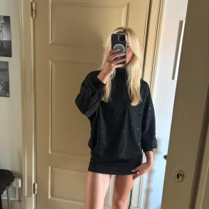 Svart glittrig kortklänning med öppen rygg - Supersnygg svart kortklänning med glittriga detaljer och öppen rygg. Klänningen har en loose passform upptill, lång ärm och hög hals. Perfekt för dig som vill sticka ut med en cool och trendig look. Materialet känns mjukt och följsamt mot kroppen.