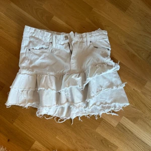 Vit jeanskjol med volanger - Säljer en vit jeanskjol med coola volanger och råa kanter. Kjolen har fickor fram och dragkedja med knapp. Perfekt för dig som gillar en edgy och trendig stil. Passar till sneakers eller boots för en chill look.