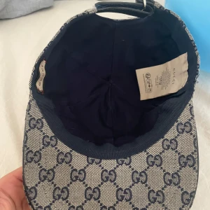 Gucci keps med GG-mönster och läderdetalj - Snygg keps från Gucci i beige och marinblått med klassiskt GG-monogram över hela kepsen. Justerbar rem bak i svart läder och ikonisk röd-blå rand på sidan. Tillverkad i bomullscanvas med exklusiv känsla och perfekt passform.