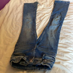 Blå bootcut jeans från H&M - Säljer ett par blå bootcut jeans från H&M med låg midja och justerbar midja. Klassisk femficksmodell med snygga detaljer på bakfickorna och slitningar framtill. Perfekta för dig som gillar en avslappnad och trendig look.