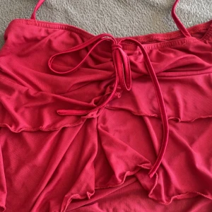 Röd topp med volang och snörning - Snygg röd topp med tunna axelband och volangdetaljer framtill. Toppen har snörning i mitten för en extra touch och en feminin look. Materialet känns mjukt och stretchigt, perfekt för stranden eller poolen. Helt ny