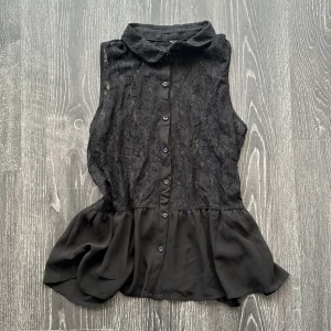 Svart ärmlös blus med spets och volang - Snygg svart ärmlös blus med spets upptill och knappar hela vägen framtill. Krage och volang nertill ger en extra touch. Perfekt för dig som gillar detaljer och en lite romantisk stil.