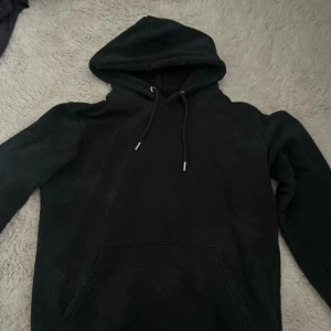 Svart hoodie från Stanley/Stella - Svart hoodie från Stanley/Stella med klassisk huva och snörning. Tröjan har känguruficka framtill och är tillverkad i ekologisk bomull och återvunnen polyester. Perfekt för chill dagar eller när du vill ha en enkel och stilren look.