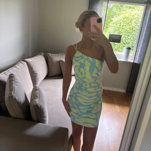 Blå och ljusgrön oneshoulder kortklänning - Säljer en superfin kort klänning med blå och ljusgrönt mönster. Klänningen har oneshoulder-design med tunna axelband och ärmlös stil. Sitter tight och har en öppen rygg, perfekt för dig som gillar att sticka ut med färg och form.