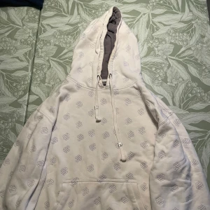 Vit hoodie från Rocawear med brodyr - Vit hoodie från Rocawear med all-over brodyr och broderad logga på ärmen. Tröjan har stor huva med snörning och känguruficka framtill. Tillverkad i bomull och polyester, perfekt för en chill streetwear-look.