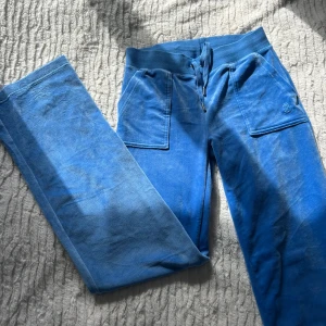 Blå velourbyxor Juicy Couture S - Blåa mjukisbyxor från Juicy Couture i mjuk velour med bred resår och snörning i midjan. Byxorna har raka ben, stora fickor fram och broderad JC-logga på ena fickan. Perfekta för en chill och trendig look.