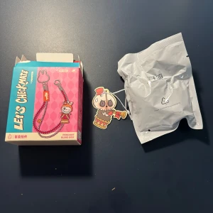 Let's Checkmate blå pawn pendant blindbox - Unikt halsband från Let's Checkmate-serien med en blå snodd och silverkedja. Hängsmycket föreställer en schackpjäs 'pawn' med en söt figur i rött och beige, som har en trumpet och cylinderhatt. Perfekt för dig som gillar schack och coola accessoarer. Labubu