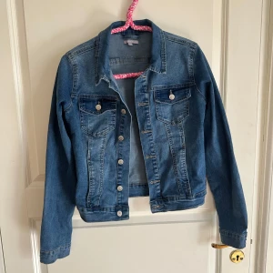 Blå jeansjacka från  - Klassisk blå jeansjacka från Sincerely Love med snygga silvriga knappar och två bröstfickor. Jackan har en rak passform, krage och långa ärmar. Perfekt att slänga över en hoodie eller t-shirt för en chill look.