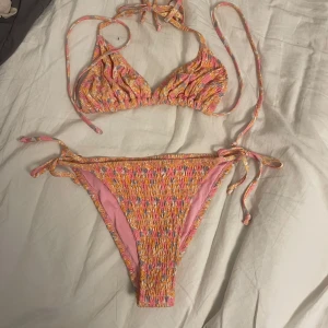 Färgglad bikini med smock och knyt - Superfin bikini i rosa, orange och gul med smockat tyg och knyt i sidorna och nacken. Triangeltopp och klassiska bikinitrosor med justerbara band. Perfekt för stranden eller poolen!