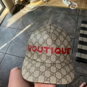 Gucci keps med GG-mönster och röd text - Snygg unik beige keps från Gucci med klassiskt GG-mönster över hela kepsen och röd text 'BOUTIQUE' framtill. Justerbar rem bak i brunt skinn med guldfärgad detalj. Perfekt för dig som vill sticka ut med lyxig streetstyle.