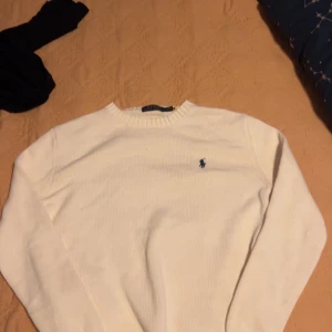 Vit stickad tröja från Polo Ralph Lauren - Vit stickad tröja från Polo Ralph Lauren med klassisk rund hals och den ikoniska blå logon broderad på bröstet. Tröjan är gjord i 100% bomull och har långa ärmar. Perfekt för dig som gillar stilrena och tidlösa plagg.