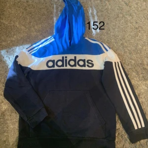 Adidas hoodie blå och vit - Säljer en snygg Adidas hoodie i blått och vitt med stor logga över bröstet och klassiska vita ränder på ärmarna. Tröjan har huva, magficka och långa ärmar. Perfekt för dig som gillar sportig stil och streetwear.