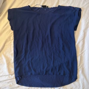 Mörkblå blus med ficka och knappdetalj - Enkel och snygg mörkblå blus med korta ärmar, rund halsringning och en liten bröstficka. Ärmarna har en cool knappdetalj på axeln och blusen är något längre baktill för en avslappnad look. Perfekt att styla med jeans eller kjol.