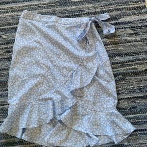 Blommig omlottkjol från Vero Moda - Supersöt ljusblå omlottkjol från Vero Moda med vita blommor och volangkant. Kjolen knyts i midjan och har en flowy känsla som passar perfekt till varma dagar. Fräsch och feminin look som är lätt att matcha med en enkel topp.