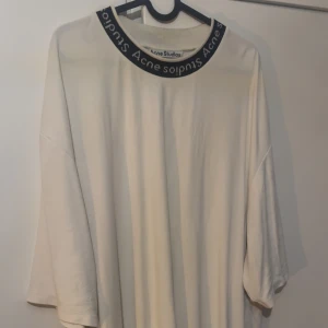 Oversized vit t-shirt Acne Studios - Vit oversized t-shirt från Acne Studios med breda, korta ärmar och rund hals med svart kant där Acne Studios-loggan är tryckt runt hela kragen. Mjuk bomullskänsla och loose fit för chill vibe.
