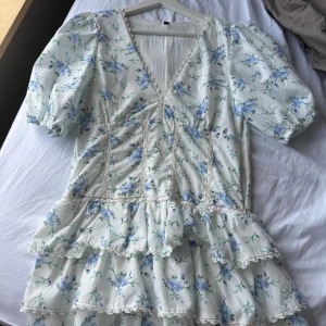 Blommig vit kortklänning med volanger - Supersöt vit kortklänning med blå blommor och gröna blad, puffärmar och v-ringning. 