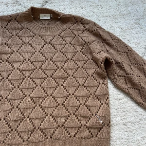Brun stickad tröja från Kaffe - Säljer en brun stickad tröja från Kaffe i storlek S/M. Tröjan har ett snyggt hålmönster med geometriska former och ribbade muddar vid hals, ärmslut och nederkant. Perfekt för dig som gillar en chill och trendig look med lite extra detaljer.