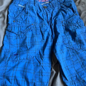 Blårutiga cargoshorts Alpha Industries - Snygga cargoshorts från Alpha Industries i klarblått med rutigt mönster. Shortsen har flera fickor med lock och knapp, dragkedja och knappstängning samt coola detaljer som logga på benet. Tillverkade i 100% bomull för en skön känsla.