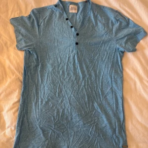 Blå t-shirt med v-ringning från EDC - Snygg ljusblå t-shirt från EDC med v-ringning och fyra svarta knappar framtill. T-shirten är kortärmad och har en chill, loose passform. Materialet är mjuk bomull, perfekt för varma dagar. Enkel och stilren design som funkar till jeans eller shorts.