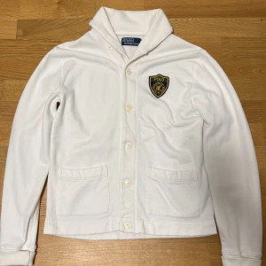 Vit overshirt från Polo Ralph Lauren - Snygg vit overshirt från Polo Ralph Lauren med klassisk krage, knäppning framtill och två fickor. På bröstet sitter en broderad Polo-patch i blått och gult. Perfekt för lager-på-lager och en clean classics look. Priset är inte fast.