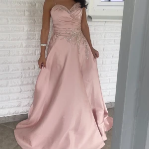 Ljusrosa långklänning med paljetter - Säljer en elegant ljusrosa långklänning med axelbandslös design och v-ringad överdel. Klänningen har vackra paljettdekorationer och draperingar över bysten samt ett glansigt satinliknande material som ger en lyxig känsla. Perfekt för bal eller fest!