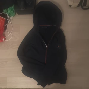 Svart cardigan från Tommy Hilfiger - Svart cardigan från Tommy Hilfiger i storlek S. Klassisk design med huva, dragkedja och diskret Hilfiger-logga på bröstet. Röd och vit kant vid halsen ger en snygg detalj. Perfekt för chill dagar och enkel att matcha med jeans eller joggers. Vid funderingar så är det bara att skriva ✍️ ✅