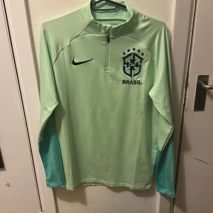 Nike Brasil långärmad fotbollströja - Snygg långärmad Brasilien fotbollströja från Nike i ljusgrön färg. Tröjan har halv dragkedja, CBF-logga och Nike Swoosh på bröstet. Ärmarna har ett diskret mönster i en mörkare grön nyans. Tillverkad i Dri-FIT material som andas.                       Passar perfekt som träningströja men även till andra sammanhang, pris går att diskutera men inga flyballs tack!:)  OBS, Detta är ett utgått plagg och ett av de få äkta som finns kvar!
