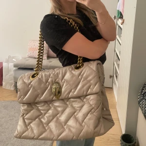 Quiltad beige axelväska Kurt Geiger - Stor quiltad axelväska från Kurt Geiger i beige skinn med chunky guldfärgad kedja och ikoniskt örnhuvud-spänne. Väskan har lyxig känsla och rymmer allt du behöver. Perfekt för dig som vill sticka ut med en statementväska. Det finns lite skav på kanterna men annars är den i super bra skick! 