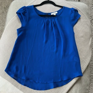 Kornblå blus storlek S - Kornblå blus från MINE i flowy polyester med rund halsringning och korta ärmar. Lätt rynk framtill och knapp i nacken. Perfekt för dig som gillar en enkel men snygg look.