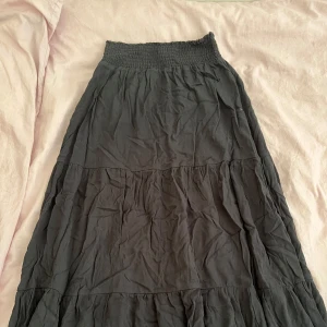 Svart långkjol med volanger - Säljer en svart långkjol med smock i midjan och flera volanglager. Kjolen är luftig och har en skön, avslappnad passform som gör den enkel att styla med olika toppar. Perfekt för dig som gillar en bohemisk vibe och vill ha något bekvämt och snyggt.