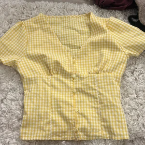 Gul rutig blus med v-ringning - Superfin gul och vit rutig blus med v-ringning och korta ärmar. Blusen har knappar framtill och är figursydd för en snygg passform. Tillverkad i mjuk polyester med lite stretch för extra komfort. Perfekt för dig som gillar färg och mönster! Köpt secondhand 