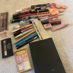Stort sminkpaket med paletter och mascara - Säljer ett stort sminkpaket med flera ögonskuggspaletter i nude och bruna toner, highlighterpalett i rosa och guld, olika mascaror, eyeliners i svart och blått, läppglans och läppstift i rosa och nude, samt sminkborste och spegel. Perfekt för dig som gillar att experimentera med olika looks.