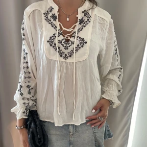 Vit broderad blus med snörning - Säljer en vit blus med svart broderi på bröstet och längs ärmarna. Blusen har snörning vid halsen och är tillverkad i ett lätt, luftigt material. 