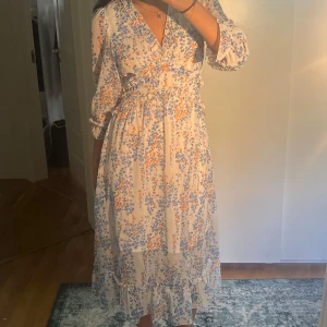 Blommig långklänning med volang - Superfin långklänning i ljus färg med blått och orange blommönster. Klänningen har omlott-v-ringning, volangdetaljer och trekvartsärmar med lätt puff. Materialet känns som tunn och luftig polyester, perfekt för varma dagar!! vet ej vart den är från då jag köpte på sellpy!