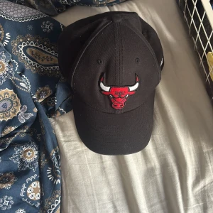 Chicago Bulls keps från New Era - Svart Chicago Bulls keps från New Era, modell 9FORTY. Broderad Bulls-logga i rött och vitt framtill, justerbar passform och klassisk böjd skärm. Tillverkad i bomull och perfekt för dig som gillar basket eller streetwear.