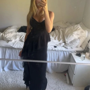 Svart långklänning med volanger och slits - Säljer en svart långklänning med smala axelband, v-ringad urringning och snygga volangdetaljer framtill. Klänningen har en hög slits på ena sidan som ger en cool och elegant vibe. Perfekt för dig som vill sticka ut med en stilren look. Storlek 32 från Gina 