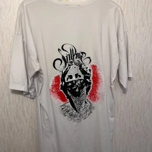 Vit t-shirt med grafiskt tryck - Vit t-shirt från Sullen i storlek XL som passar m-xxl (slash) med stort grafiskt tryck på ryggen i svart och rött. Trycket har coola detaljer och texten 'It’s called the hustle'. T-shirten har rund hals och korta ärmar, tillverkad i mjuk bomull för skön passform.