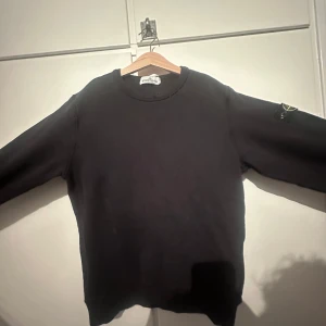 Svart Stone Island sweatshirt junior - Storlek: 156. Liten slitning på kragen därav det billiga priset. Svart sweatshirt från Stone Island Junior med klassisk rund hals och ikonisk logga på ärmen. Tröjan har en clean look och är tillverkad i mjuk bomull, perfekt för dig som gillar streetwear och vill ha något stilrent men ändå med edge.