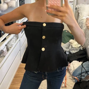 Svart bandeau tubtopp från Vero Moda - Svart bandeau tubtopp från Vero Moda med snygga guldknappar framtill. Toppen har en lätt utsvängd form nedtill och är helt axelbandslös. Perfekt för dig som vill ha en stilren och trendig look. Säljer pga att jag endast bär silver och att den är förstor, endast testad!