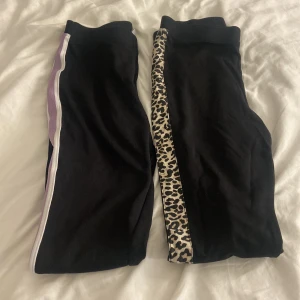 Svarta leggings med leopard och lila detaljer - Två par svarta leggings från Lindex i storlek 170. Ena paret har leopardmönstrad sidoremsa, det andra har lila och vita ränder längs benen. Båda är i stretchigt bomullsmaterial och har en tight passform som sitter snyggt på benen.