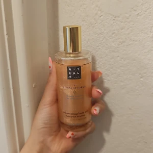 Rituals The Ritual of Karma Shimmer Oil - Shimmering body oil från Rituals The Ritual of Karma med lotus och vit te. Oljan har en guldig, skimrande finish som ger huden glow och lyster. Kommer i en genomskinlig glasflaska med guldfärgat lock. Perfekt för att boosta din hud med extra shine.