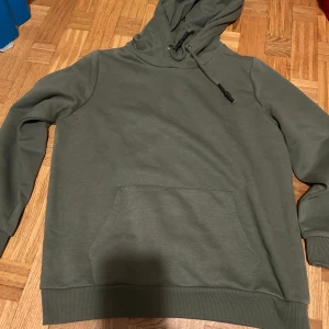 Olivgrön hoodie från Clodus - Säljer en olivgrön hoodie från Clodus i storlek L. Tröjan har en stor magficka, huva med snörning och ribbade muddar vid ärmslut och nederkant. Perfekt för chill dagar och enkel att matcha med jeans eller joggers.