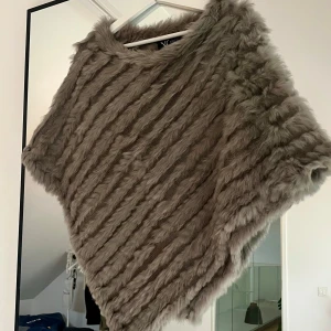 Beige pälsponcho från Nature's Collection - Supermjuk och fluffig beige poncho från Nature's Collection. Den har korta ärmar och är gjord i fuskpäls med snedställda ränder. 