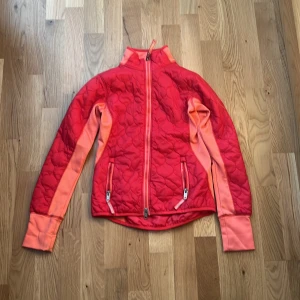 Röd quiltad jacka från Jacson - Snygg röd quiltad jacka från Jacson med orange detaljer längs ärmarna och dragkedja framtill. Jackan har hög krage, två fickor med dragkedja och coolt mönstrad yta. Perfekt för dig som gillar färg och vill sticka ut i stallet eller på stan.bra för den kommande hösten