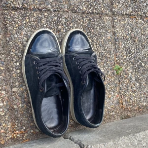 Svarta sneakers från Lanvin med lackad tå - Snygga svarta sneakers från Lanvin med klassisk rund tå och lackad tåhätta. Ovandelen är i mocka och skorna har svarta snören samt vit sula. Perfekta för dig som gillar stilrena och exklusiva detaljer i din vardag.