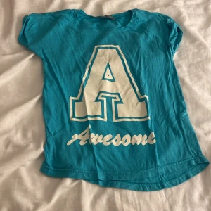 Turkos t-shirt med tryck från Lindex - Turkos t-shirt från Lindex med stort vitt A och texten 'Awesome' på framsidan. Rund hals och korta ärmar, tillverkad i mjuk bomull. 