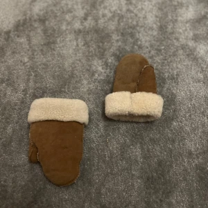Bruna mockavantar med foder - Säljer ett par bruna tumvantar i mocka med mjukt, ljust foder och breda uppvikta muddar. Perfekta för kalla dagar när du vill hålla händerna varma och mysiga. Klassisk design som funkar till det mesta. Vet ej storlek men de passar 1 åringar 