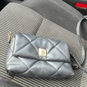 Svart quiltad axelväska i skinn - Snygg svart axelväska i quiltad design med guldfärgat spänne framtill. Väskan har en lång axelrem och är tillverkad i mjukt skinn. Perfekt storlek för det viktigaste och har flera fack inuti för bra organisering.