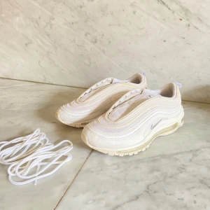 Nike Air Max 97 vita sneakers - Säljer ett par fräscha Nike Air Max 97 i helvitt med diskreta silverdetaljer och synlig luftbubbla i sulan. Skorna har en vågig design, rund tå och kommer med extra vita skosnören. Perfekta för dig som gillar stilrena sneakers.