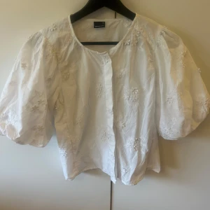 Vit blus med puffärmar från Gina Tricot - Säljer en söt vit blus från Gina Tricot med broderade blommor och puffiga korta ärmar. Blusen har rund halsringning och knäppning framtill. Perfekt för en fräsch och somrig look! Storlek S